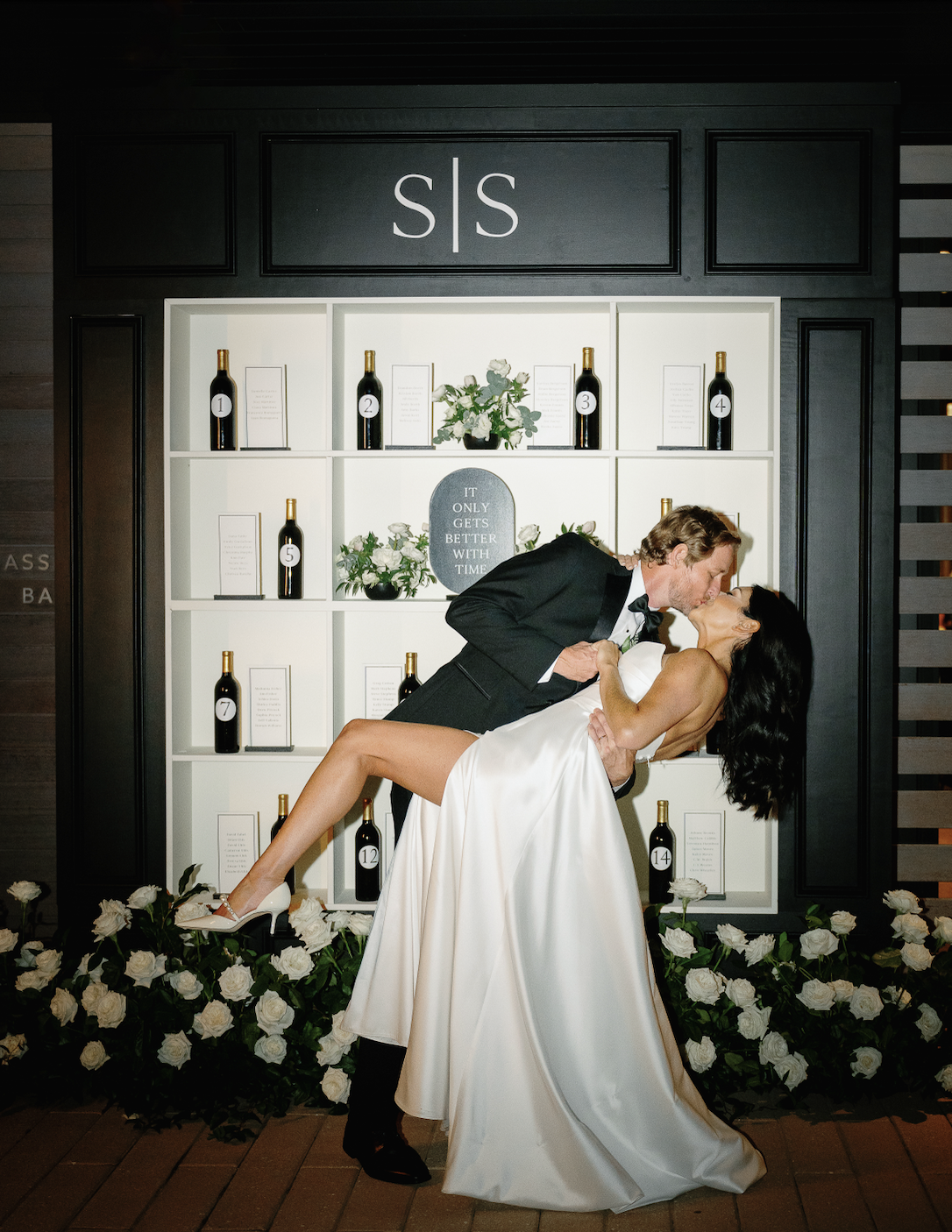 A couple shares a kiss in front of a wall with wine bottles and initials "S S." The woman wears a white dress, and the man wears a suit. White flowers decorate the scene.